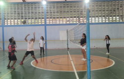 #paratodosverem: em uma quadra poliesportiva, 9 alunos jogam voleibol. À esquerda da rede, é possível ver 5 jovens e à direita, 4. Um dos jovens à esquerda está com as mãos levantadas para receber e jogar a bola que está no ar, em sua direção.