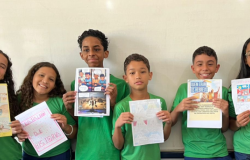 #paratodosverem: na imagem, um grupo de crianças posa sorridente em frente a um quadro branco, exibindo trabalhos de História. Elas estão em pé e vestem uniforme escolar e seguram produções coloridas com textos, desenhos e histórias em quadrinhos sobre povos antigos. 