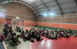 Na quadra de uma escola, seis filas de crianças sentadas com mochilas ao lado aguardam a saída, interagindo entre si. O espaço tem chão colorido, arquibancada verde, parede laranja, tabela de basquete, trave com rede e claraboias no teto que iluminam o ambiente.