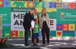 #paratodosverem: na imagem, o Prefeito e os(as) secretários(as), trajados(as) formalmente, posam junto a um aluno da RME-BH para uma foto, em primeiro plano. Ao fundo, há um banner, referente à Semana do Meio Ambiente, contendo elementos coloridos que representam a fauna e a flora.