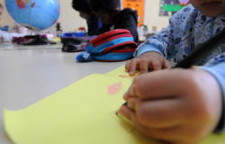 #paratodosverem: a foto mostra crianças em uma sala de aula envolvidas em atividades escolares. Em primeiro plano, uma criança segura um lápis preto e desenha em uma folha amarela, com um traço visível em vermelho. Ela veste uma blusa azul com estampas. Ao fundo, outras crianças estão concentradas em suas tarefas, e, sobre a mesa, há materiais escolares, como estojo e um globo terrestre.