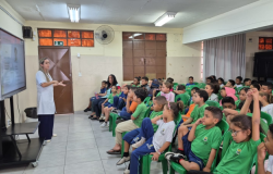 #paratodosverem: sala de aula com fileiras de cadeiras verdes ocupadas por crianças em uniforme escolar, atentas a uma apresentação. À frente, uma mulher de jaleco branco fala e gesticula, ao lado de uma tela digital ligada. O espaço tem paredes claras, janelas com vidros alaranjados e cortina cinza ao fundo.