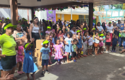 #paratodosverem: a foto mostra uma confraternização com crianças pequenas e pessoas adultas reunidas em um momento coletivo de evento comemorativo ao ar livre. As crianças, muitas usando viseiras verdes, acompanham uma apresentação. 