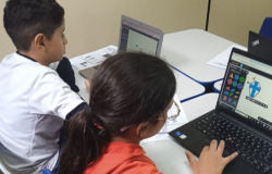 #paratodosverem: Dois estudantes estão sentados lado a lado em uma mesa, cada um usando um notebook. O menino à esquerda, de camiseta branca, olha para a tela. A menina à direita, de óculos e camiseta vermelha, interage com um programa de formas geométricas coloridas. Há folhas de papel sobre a mesa e ao fundo uma parede clara.