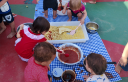 #paratodosverem: crianças pequenas brincam sentadas no chão, sobre toalha xadrez branco com azul. Elas manipulam bandejas com macarrão cozido e tigelas com molho vermelho, usando colheres e as mãos. A atividade acontece ao ar livre, em piso colorido de verde e vermelho. Algumas crianças estão descalças e vestidas com roupas leves e coloridas.
