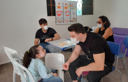 #paratodosverem: em uma sala de paredes brancas, um jovem de camiseta preta e máscara conversa com uma estudante de cabelo preso e blusa azul, que está sentada em frente a ele, que segura um palito de madeira. Ao fundo, estão sentados(as) outro jovem e uma mulher, ambos de máscara. Na parede, há um cartaz pendurado na janela, ilustrando cuidados bucais.