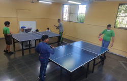 #paratodosverem: a foto mostra uma sala, com paredes amarelas e amplas janelas gradeadas. No centro da sala, há duas mesas de tênis de mesa onde quatro alunos estão envolvidos na atividade esportiva, com raquetes nas mãos e atentos ao jogo. As crianças vestem uniformes escolares ou roupas esportivas. Ao fundo, duas crianças estão sentadas próximo à parede.