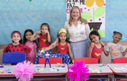 #paratodosverem: na foto, oito meninas fantasiadas de super-heroínas e uma professora posam sorridentes atrás de mesas com trabalhos manuais coloridos, em uma sala de aula. Atrás da professora, há um quadro decorado de azul claro e moldura de papel marrom.