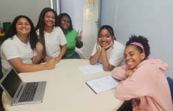 #paratodosverem: na imagem, cinco estudantes estão sentadas em torno de uma mesa redonda, sorrindo para a câmera. Três usam camisetas brancas com o brasão da Prefeitura de Belo Horizonte; outra está com camisa de uniforme verde contendo o brasão, e a quinta veste moletom rosa. Sobre a mesa, há um notebook e cadernos abertos. Ao fundo, há uma parede azul-clara, uma porta branca e um cartaz com a palavra “PAS”.