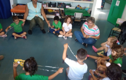 #paratodosverem: na imagem, tirada em uma perspectiva de cima para baixo, diversas crianças uniformizadas estão sentadas em círculo, no chão de uma sala. Entre elas, há três mulheres adultas. Atrás delas, estão três prateleiras brancas cheias de brinquedos, algumas cadeiras e duas mesas pequenas (uma com toalha alaranjada e outra com toalha verde). No canto superior direito, há uma porta aberta com o número nove estampado.