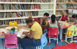 #paratodosverem: na imagem, há uma biblioteca com estantes bege repletas de livros de diversas cores e com algumas bonecas. Em foco, estão duas mesas coloridas, que apoiam as sacolas. Sobre as mesas, também há papéis, pincéis e tintas de diversas cores. Ao redor das mesas, sentadas em cadeiras coloridas, funcionárias da escola em trajes diversos pintam sacolas. 