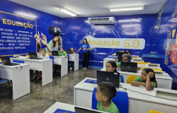 #paratodosverem: fotografia de sala de aula com paredes azuis em que está escrito “Educação 5.0”. Há várias crianças uniformizadas sentadas, sozinhas ou em duplas, mexendo em computadores, com uma educadora auxiliando-as.