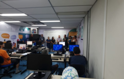 #paratodosverem: em uma sala equipada com computadores, seis estudantes estão sentados(as) em suas mesas, atentos(as) a oito formadores(as) que estão em pé, sobre um piso azul, à frente da sala. Atrás destes(as), está afixado um quadro branco, e, nas paredes laterais, há uma decoração colorida com palavras como “tecnologia” e “cidadão”. 