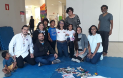 #paratodosverem: um grupo de pessoas adultas e crianças posa para foto, sobre um tatame azul, em um espaço interno. Sobre o tatame, há folhas de papel, fotos, tesouras e caixas de lápis. Algumas pessoas estão sentadas, outras de pé. Ao fundo, paredes brancas e colunas, com cartazes coloridos.