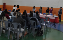 #paratodosverem: na imagem, pessoas sentadas em cadeiras cinzas acompanham uma apresentação, em um ginásio com paredes pintadas nas cores laranja e branco. À frente, seis pessoas estão em pé, próximas a uma mesa com toalha rosa e vasos de flores, e uma mulher segura o microfone. No canto direito, um projetor sobre tripé aponta para a parede.