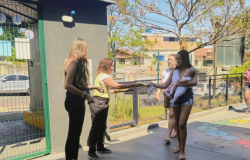 #paratodosverem: Uma mulher de blusa amarela e calça preta entrega um panfleto a uma jovem de camiseta branca sem mangas e shorts jeans, que segura um bebê no colo. Ao lado, outra mulher de preto observa. Atrás, há uma senhora de blusa branca e saia estampada. A cena ocorre em área externa ensolarada, com muros, árvores e casas ao fundo.