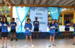 #paratodosverem: A imagem mostra um grupo de crianças se apresentando em frente a um painel azul com os dizeres “Setembro Azul 26/09 Dia Nacional dos Surdos”. Elas vestem camisetas azuis, shorts ou saias da mesma cor e usam luvas brancas, posicionadas em gestos que sugerem linguagem de sinais. Ao fundo, há cortinas azuis presas a uma estrutura amarela.