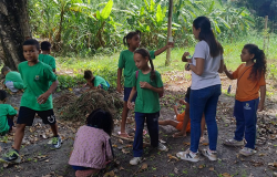 #paratodosverem: a foto mostra um grupo de estudantes e uma educadora em área ao ar livre, com vegetação abundante ao fundo, incluindo bananeiras. As crianças vestem uniformes escolares em cores verde e laranja. No centro da imagem, três delas interagem com a educadora, enquanto outras estão ajoelhadas na terra, sentadas ou em pé.
