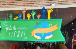 #paratodosverem: na foto, há um painel verde, em primeiro plano, com moldura brilhante, que exibe a frase “Festa da Família”, a mensagem “Salve o Planeta” sobre um desenho da Terra e mãos segurando-a. Acima, mãos coloridas nas cores verde, azul, marrom e amarelo, com rostos sorridentes, completam a decoração. Ao fundo, pessoas conversam em um corredor decorado com murais.