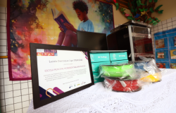 Escola recebe o kit de impressora 3D como destaque da Liga STEAM 2025. #paratodosverem: A imagem mostra uma mesa coberta com toalha branca, onde estão dispostos um certificado de destaque da Liga STEAM e caixas com materiais coloridos de filamentos para impressora 3D. Ao fundo, há um painel ilustrado com a imagem de uma criança lendo um livro.