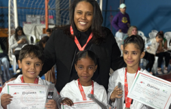 #paratodosverem: em um ginásio esportivo, uma mulher vestida de preto sorri enquanto abraça três crianças que seguram diplomas e medalhas. As crianças vestem uniformes brancos de taekwondo e usam medalhas vermelhas no pescoço. A mulher também usa uma medalha semelhante. Ao fundo, há cadeiras brancas ocupadas por outras pessoas e uma quadra com rede de futsal. O clima é de comemoração.