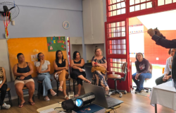#paratodosverem: a foto mostra uma roda de conversa em uma sala de aula, onde um grupo de pais e responsáveis assiste à apresentação de Telly Almeida. Ele está em pé, gesticulando. Um projetor está ligado sobre uma mesa, ao lado de um notebook, no centro da sala. Ao fundo, há um cartaz com a frase "Feliz Aniversário" e desenhos infantis afixados em uma parede laranja.