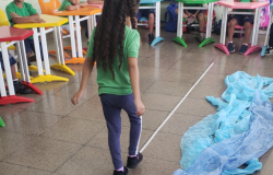 #paratodosverem: Em sala de aula, uma menina caminha sobre uma linha branca no chão de uma sala, enquanto outras crianças, sentadas em carteiras coloridas, observam. Ao lado da linha, há um tecido azul amontoado no chão. Ao fundo, vêem-se mochilas penduradas na parede