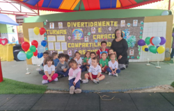 #paratodosverem: A foto mostra um grupo de crianças sentadas no chão em frente a um mural decorado, acompanhadas por uma mulher em pé ao lado direito. O mural tem o tema “Divertidamente e Caracol Compartilhando as Emoções”, com letras grandes em amarelo, desenhos coloridos, mãos pintadas e fotos das crianças coladas. Em volta, há balões coloridos em suportes brancos. A cena acontece sob uma cobertura de lona colorida.