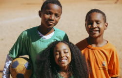 #paratodosverem: Em um close-up, duas crianças, um menino de uniforme verde e um de uniforme laranja, olham sorrindo para a câmera. Entre eles, em primeiro plano, uma menina com cabelos cacheados e volumosos sorri de olhos fechados. O menino de verde segura uma bola de futebol na lateral. O sol ilumina os rostos do grupo, transmitindo alegria e conexão.