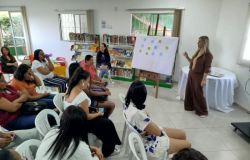 #paratodosverem: a imagem mostra uma sala pintada de branco, com estantes repletas de livros entre duas janelas gradeadas ao fundo. Sentadas em cadeiras brancas de plástico, pessoas adultas estão atentas a uma mulher em pé, ao lado de um cavalete que sustenta um quadro com post-its verdes e azuis. Atrás da palestrante, há uma mesa redonda coberta por toalha branca.