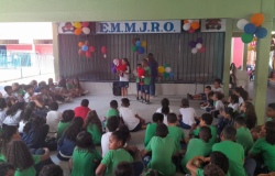 #paratodosverem: a imagem mostra um grupo de estudantes reunidos(as) em espaço coberto da EM Monsenhor João Rodrigues de Oliveira. Três crianças, com fantoches, apresentam-se à frente dos(as) colegas, acompanhadas por uma professora. Balões coloridos decoram o local. Os(As) demais alunos(as) assistem à apresentação sentados(as).