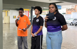 #paratodosverem: em uma área semiaberta da escola, três estudantes estão de pé, um garoto, à esquerda, e duas garotas, à direita. O menino segura um microfone enquanto lê seu discurso em um papel que segura, enquanto as meninas aguardam o momento de falar.