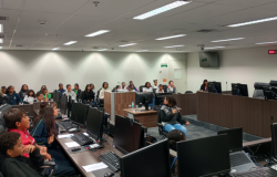 #paratodosverem: A imagem mostra uma sala ampla com mesas em formato de “U” equipadas com computadores e microfones. Diversas crianças e adolescentes estão sentados nas cadeiras, alguns na parte central e outros nas laterais, enquanto uma menina segura um microfone. Ao fundo, há mais pessoas sentadas em fileiras. O ambiente é um plenário moderno.
