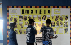 #paratodosverem: Três jovens estão de frente para um grande mural de azulejos, com o título "Amarelo" em destaque. O mural exibe recortes de cabeças de várias pessoas sorrindo e textos inspirados no álbum de Emicida. Os jovens, de costas, estão concentrados, lendo o conteúdo e interagindo com a arte em um corredor escolar.
