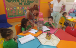 #paratodosverem: A imagem mostra uma sala de aula colorida com mesas e cadeiras infantis. Quatro crianças com camisetas verdes estão sentadas desenhando em cadernos, enquanto uma professora mostra as borboletas dentro de um pote transparente. Outra mulher observa em pé. Ao fundo, há um mural com flores e a frase “Nascem das sementes de hoje!”