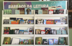 #paratodosverem: A imagem mostra uma estante branca com várias prateleiras repletas de livros de diferentes tamanhos e cores. Na parte superior há uma faixa com o título “Barraco da Leitura” em letras grandes brancas sobre um fundo colorido. Entre os livros, é possível ver obras literárias infantis e juvenis, além de alguns títulos voltados à arte e cultura afro-brasileira. O ambiente é iluminado e tem uma parede verde clara ao fundo.