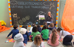 #paratodosverem: Em um espaço escolar decorado com um grande quadro preto, uma professora de blusa cinza lê um livro para um grupo de crianças pequenas, sentadas no chão. O quadro exibe a frase "BIBLIOTECANDO - LEITURAS QUE TRANSFORMAM". A cena, de fundo laranja vibrante, transmite um momento de contação de histórias e incentivo à leitura.