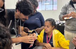 #paratodosverem: Duas mulheres e dois homens adultos, além de uma estudante de jaqueta amarela, estão reunidos em torno de uma mesa, em um close-up. O grupo examina pequenos pedaços de papel. A estudante de jaqueta amarela e um dos homens olham atentamente para os papéis nas mãos. O ambiente é um espaço interno.