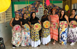 #paratodosverem: A imagem mostra oito mulheres sorridentes em uma apresentação cultural. Elas vestem saias longas coloridas e seguram peneiras decoradas com flores. Ao fundo, há um painel com tecidos estampados
