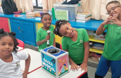 #paratodosverem: Quatro crianças estão em uma sala de aula, sorrindo em volta de uma caixa colorida com o nome “Caixa Musical”. Duas delas vestem camisetas verdes e duas, brancas, todas com o logotipo da Prefeitura de Belo Horizonte. O ambiente é alegre, com mesas, cadeiras vermelhas e prateleiras azuis cheias de livros e materiais escolares.