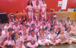 #paratodosverem: A imagem mostra um grande grupo de crianças sentadas no chão, todas vestidas com roupas brancas e camisetas com o escrito “Belezas de Minas Gerais”. Atrás delas, quatro adultos também vestidos de branco seguram instrumentos de capoeira, como berimbau e pandeiro. O fundo tem uma parede vermelha com o título “Belezas de Minas Gerais” e uma mesa coberta com tecido colorido e objetos decorativos.