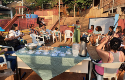 #paratodosverem: A imagem mostra um encontro de pais ao ar livre, com participantes sentados em círculo enquanto uma mulher conduz a conversa. Há uma mesa central com lanche, copos e uma garrafa térmica.