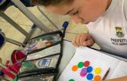 #paratodosverem: Vista superior de um estudante sentado à sua mesa escolar, vestindo uma camiseta branca com o brasão da Prefeitura de Belo Horizonte. Sobre um caderno pautado, há diversas fichas circulares coloridas (verdes, laranjas, azuis, vermelhas, amarelas e roxas) e um lápis. Ao lado do caderno, um estojo aberto revela lápis de cor e outros materiais escolares.