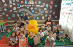 #paratodosverem: Em uma sala decorada com a frase "Estrada Literária", um grupo de crianças pequenas está sentado no chão com suas professoras. As crianças seguram sacolas de papel pardo decoradas com estrelas coloridas. O cenário é festivo, com confetes e um fundo que simula uma estrada com livros. 