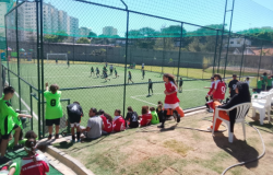 #paratodosverem: Crianças assistem e participam de um jogo de futebol em um campo cercado por tela. Alguns jogadores estão em campo, outros, de uniforme vermelho ou verde, aguardam do lado de fora. Há pessoas sentadas em cadeiras plásticas brancas, enquanto prédios e árvores aparecem ao fundo sob um céu azul.