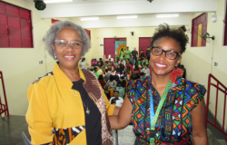 #paratodosverem: Duas mulheres sorriem para a câmera em auditório cheio de estudantes ao fundo. Ambas usam roupas coloridas com estampas afro-brasileiras. A mulher à esquerda tem cabelos grisalhos curtos e veste uma blusa amarela com detalhes estampados; a mulher à direita usa óculos, brincos grandes vermelhos e um vestido estampado.