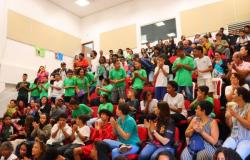 #paratodosverem: Interior de um auditório com degraus e poltronas vermelhas ocupadas por um grande grupo de jovens e adultos. Muitos estudantes vestem camisetas verdes de uniforme e aparecem aplaudindo. As paredes são brancas com painéis retangulares claros. A iluminação vem de luminárias quadradas no teto.