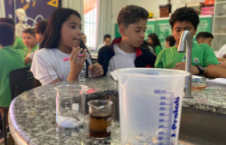 #paratodosverem: A imagem mostra estudantes participando de uma atividade científica em um laboratório escolar. Eles observam atentamente materiais como béqueres e um copo medidor sobre a bancada de granito, enquanto discutem e fazem anotações. 
