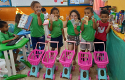 #paratodosverem: A imagem mostra sete crianças em uma sala de aula colorida. Seis delas estão em pé, sorrindo e segurando notas de dinheiro de brinquedo, e uma está sentada digitando em um teclado. À frente delas há quatro carrinhos de compras de brinquedo rosa e azul. Ao fundo, sobre uma mesa coberta por um tecido verde, há diversos produtos, como xampus, sabonetes e cremes, simulando um mercado. As crianças vestem uniformes verdes da rede municipal e demonstram alegria durante a atividade.