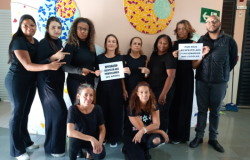 #paratodosverem: Um grupo de dez funcionários escolares, em sua maioria mulheres, está posando em frente a uma parede decorada com mosaicos. Eles vestem roupas pretas e seguram cartazes com mensagens de protesto, como "Operação Respeito aos Professores (Eu Apoio)”.